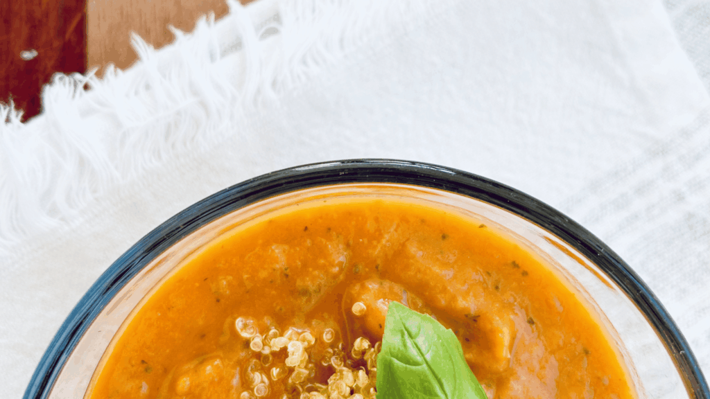 Bowl of cozy blended tomato vegetable soup garnished with roasted quinoa and a fresh basil leaf